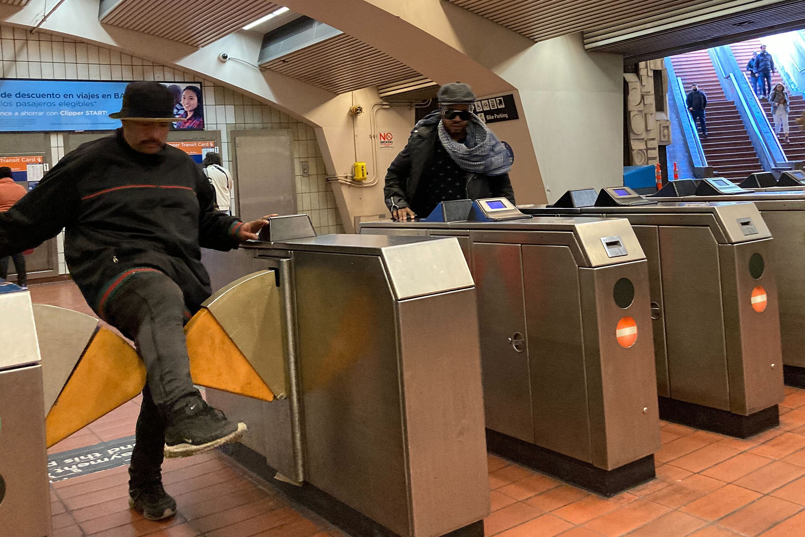 Man in black sweater steps over an orange fare gate in a train station