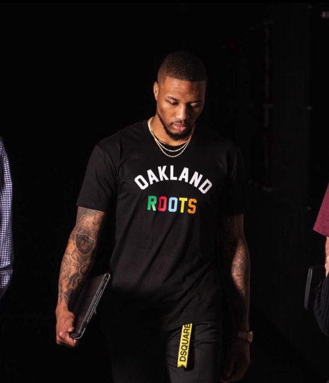 A man with tattoos wearing a black "Oakland Roots" shirt holds a laptop, walking purposefully.