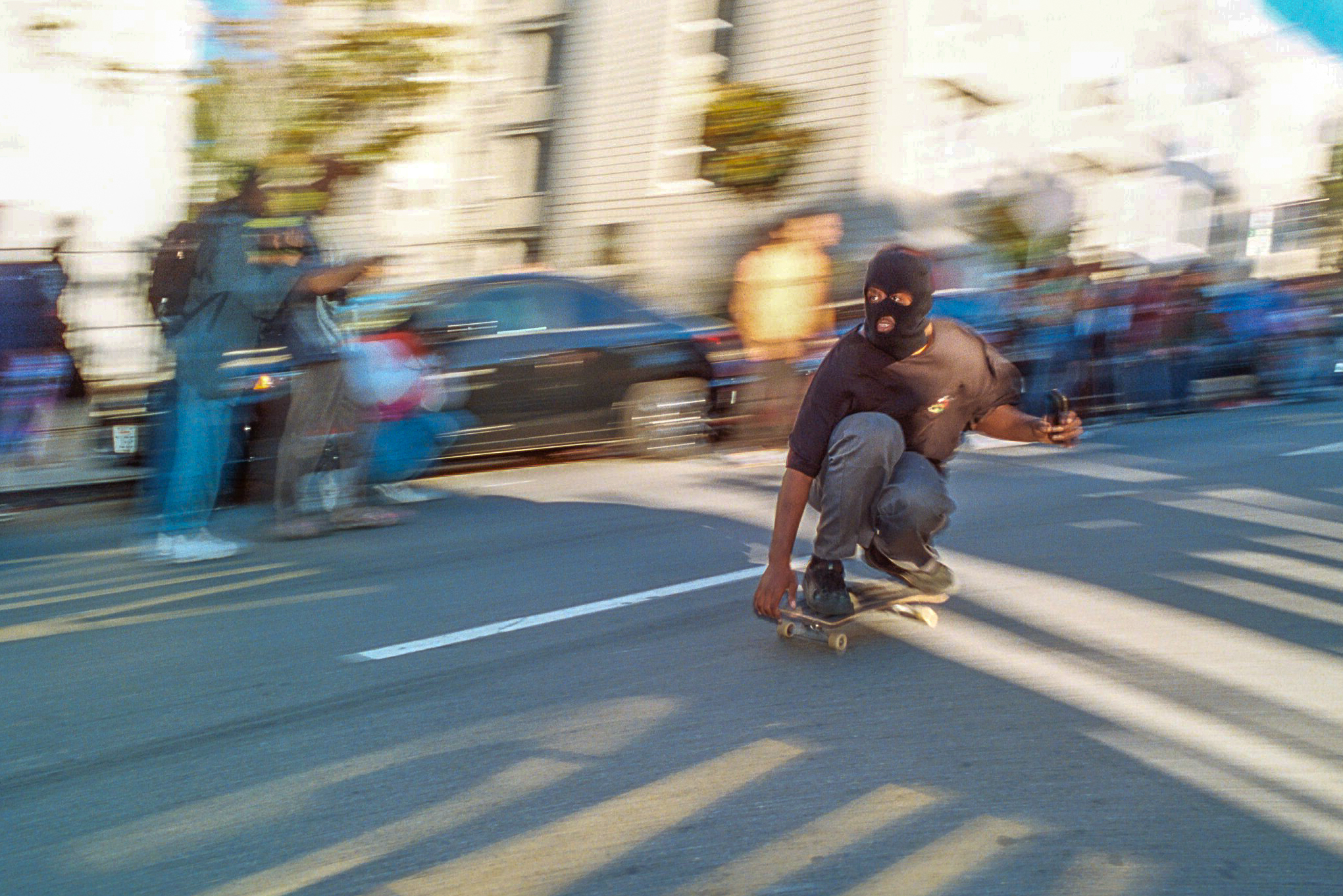 A person in a ski mask speeds on a skateboard on a bustling street.