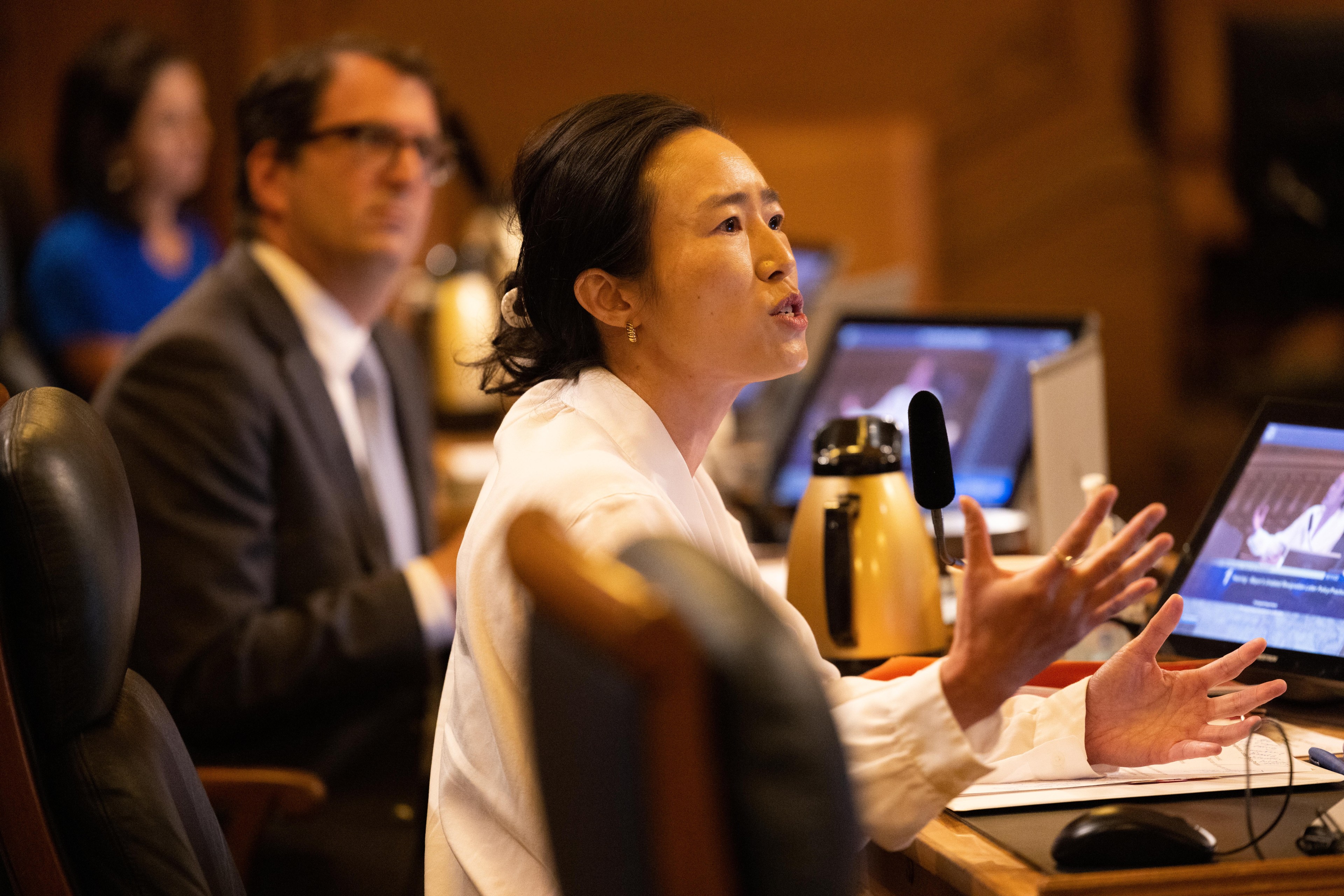 Supervisor Connie Chan, wearing a white top, looks exasperated and gestures with her hands while speaking in the Board of Supervisors chambers at City Hall. 