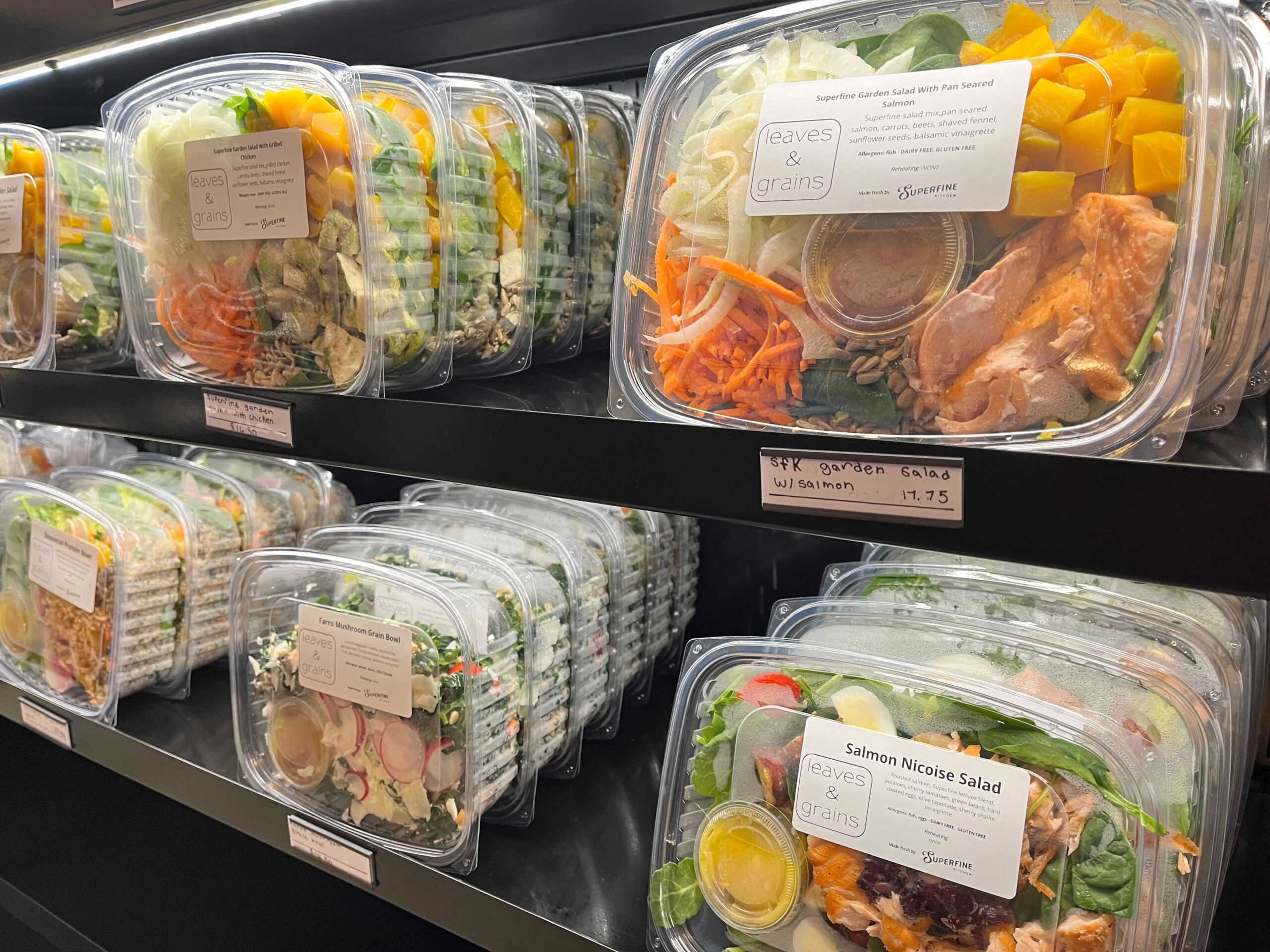 The image shows shelves filled with pre-packaged salads in clear plastic containers, each labeled with salad names and prices, featuring vibrant ingredients like salmon.