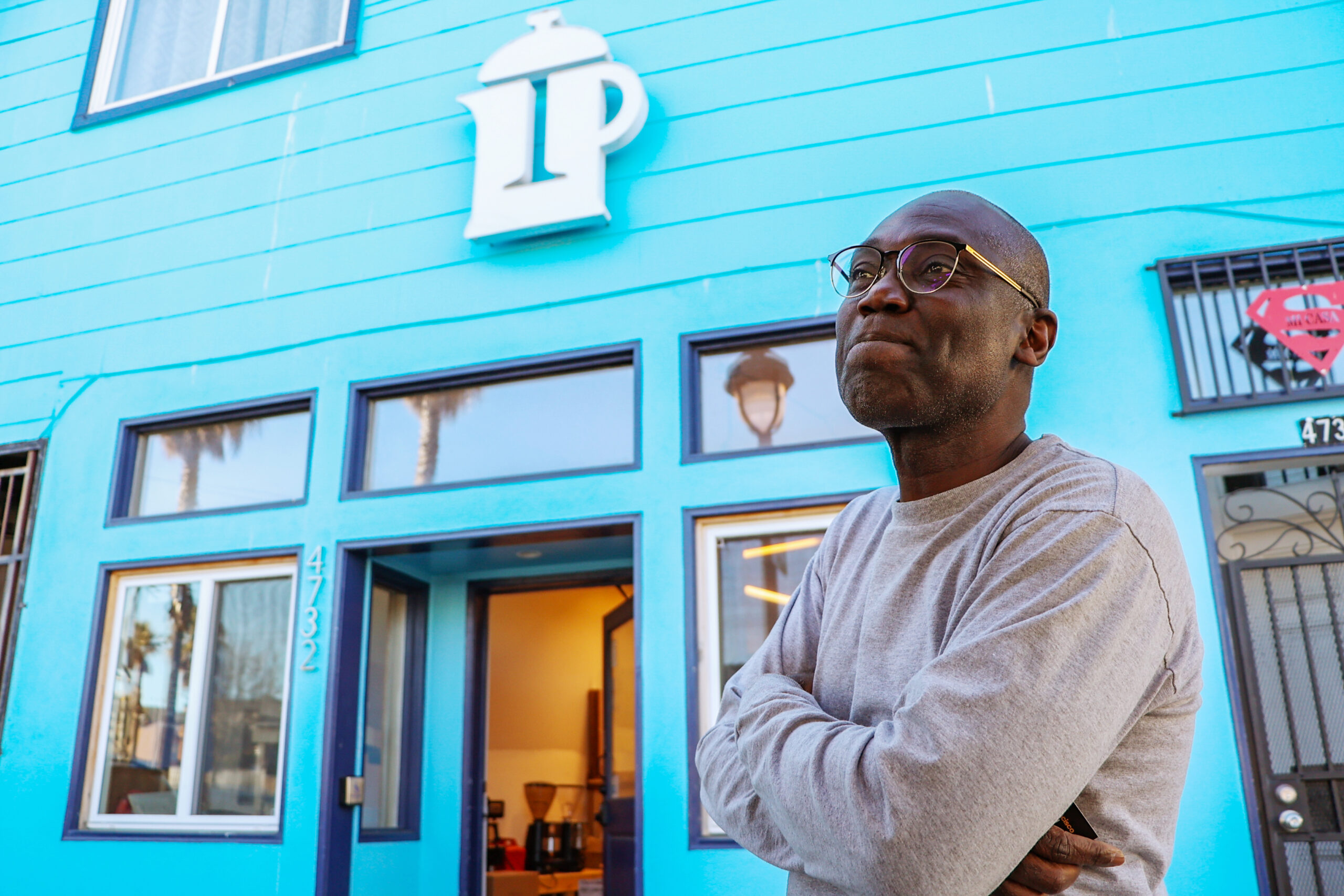 A person in glasses stands with crossed arms in front of a bright blue building featuring a coffee pot logo and the number 4732 by the door.