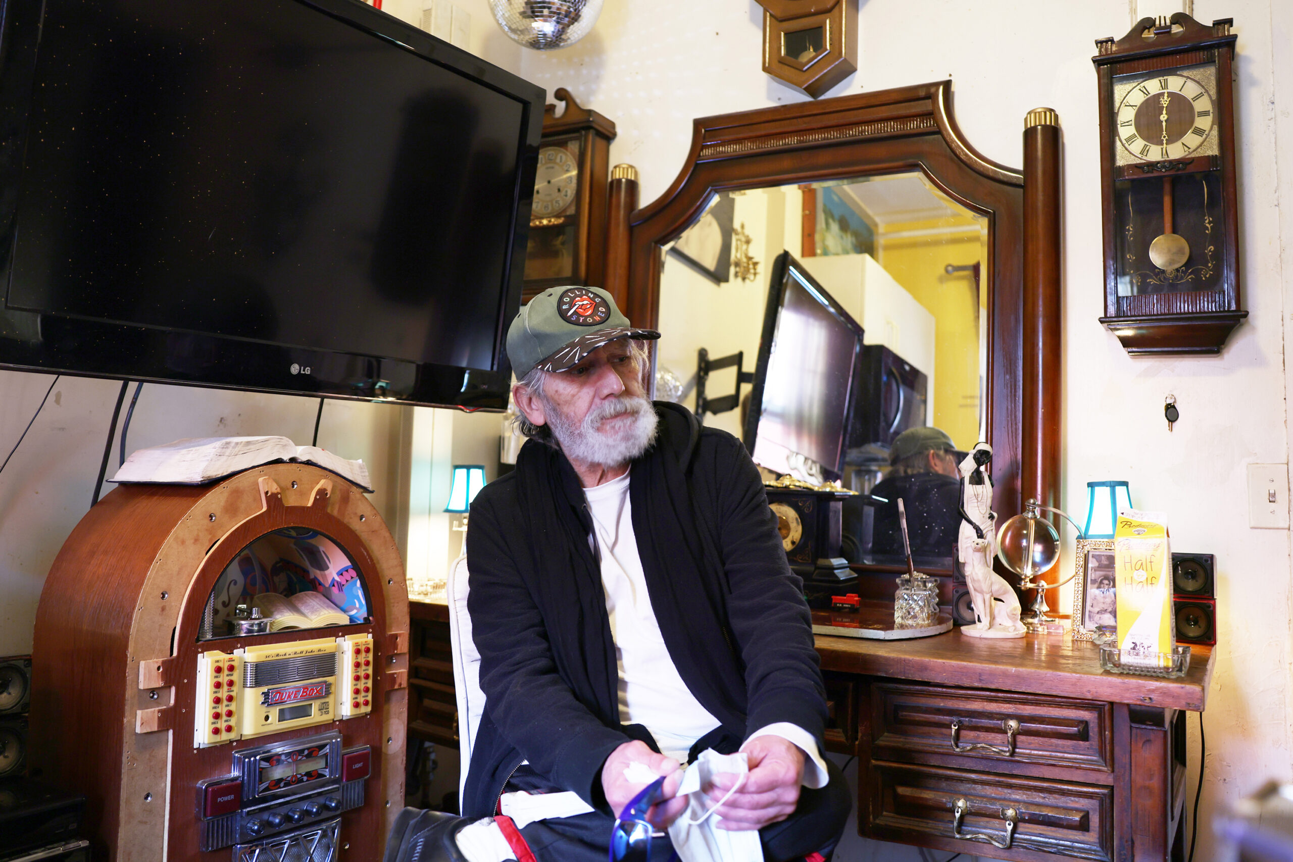 An elderly man with a beard sits by vintage items, including a jukebox, old clocks, and a mirror reflecting the room.