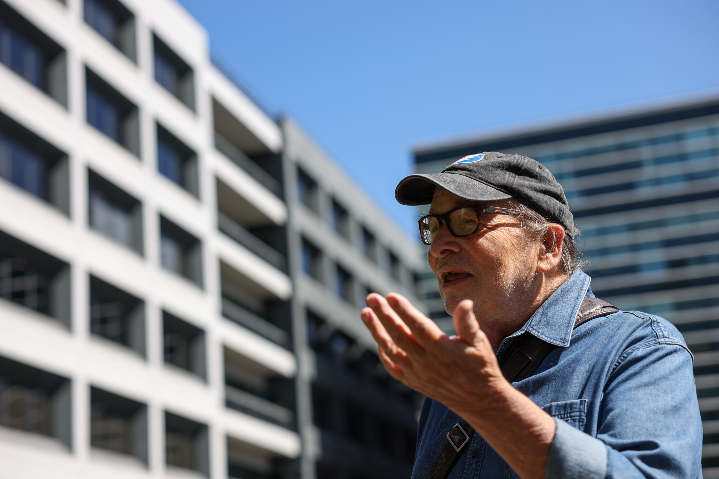 An elderly man in a cap, gesturing with his hands, with blurry buildings in the background.