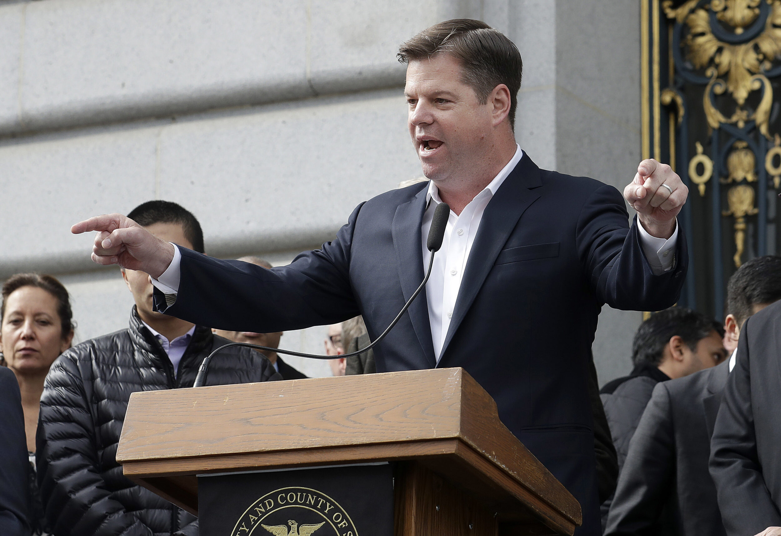 A man in a suit gestures emphatically at a podium with microphones, flanked by onlookers.