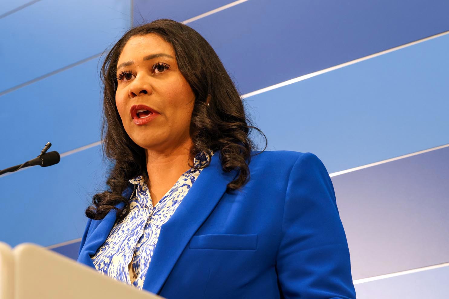 A woman in a blue jacket stands behind a podium speaking.