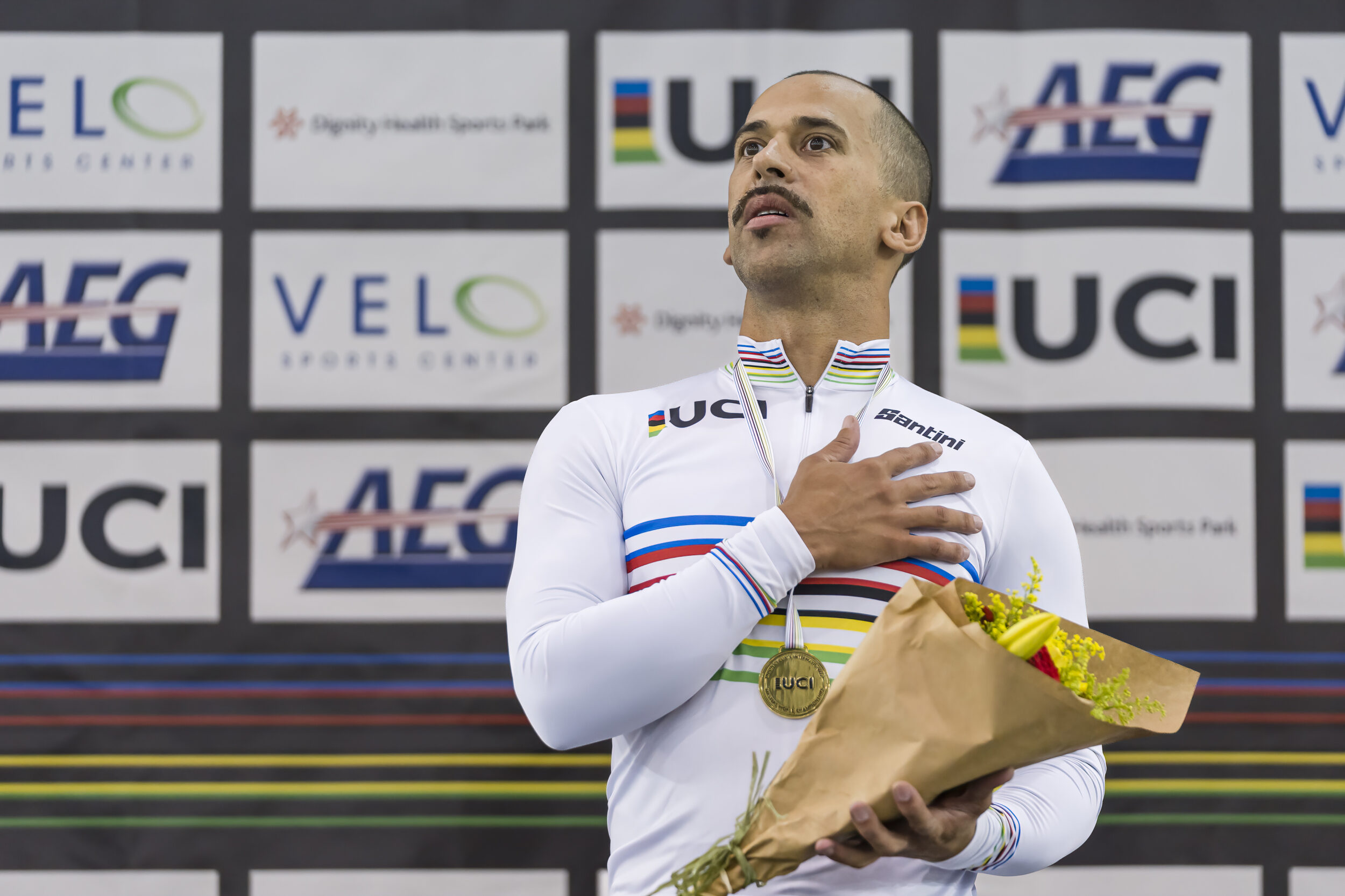 A man in a white cycling jersey and gold medal around his neck holds his right hand over his heart while clutching a bouquet of flowers in the other.