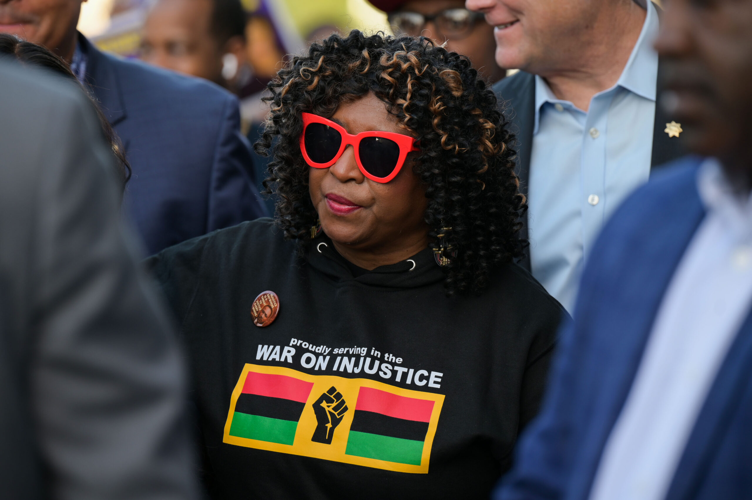 A person with red glasses, curly hair, wearing a "War on Injustice" shirt, stands among others in a gathering.