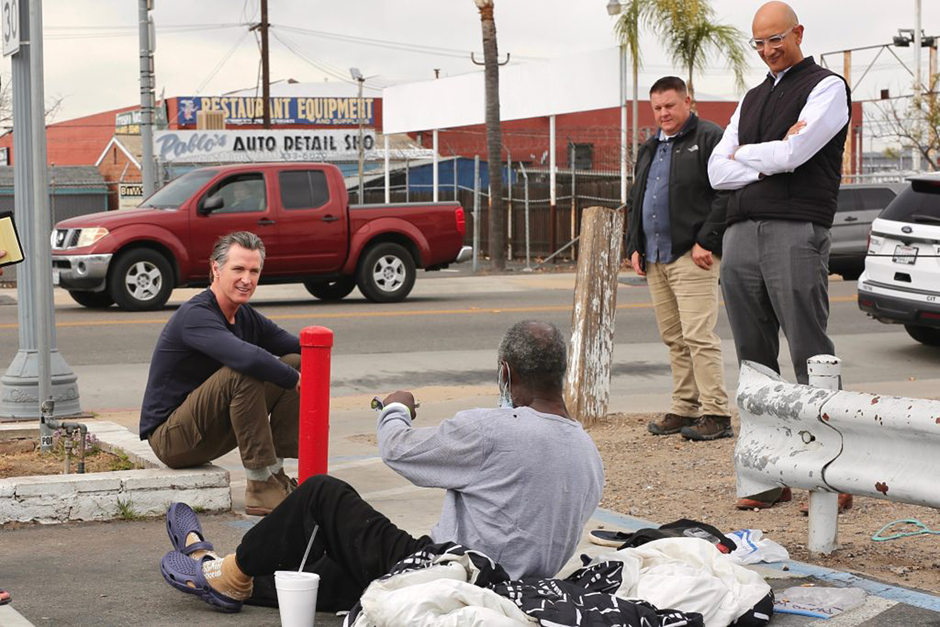 White man sits on sidewalk while speaking to Black man sitting on pavement as two other men stand nearby looking on