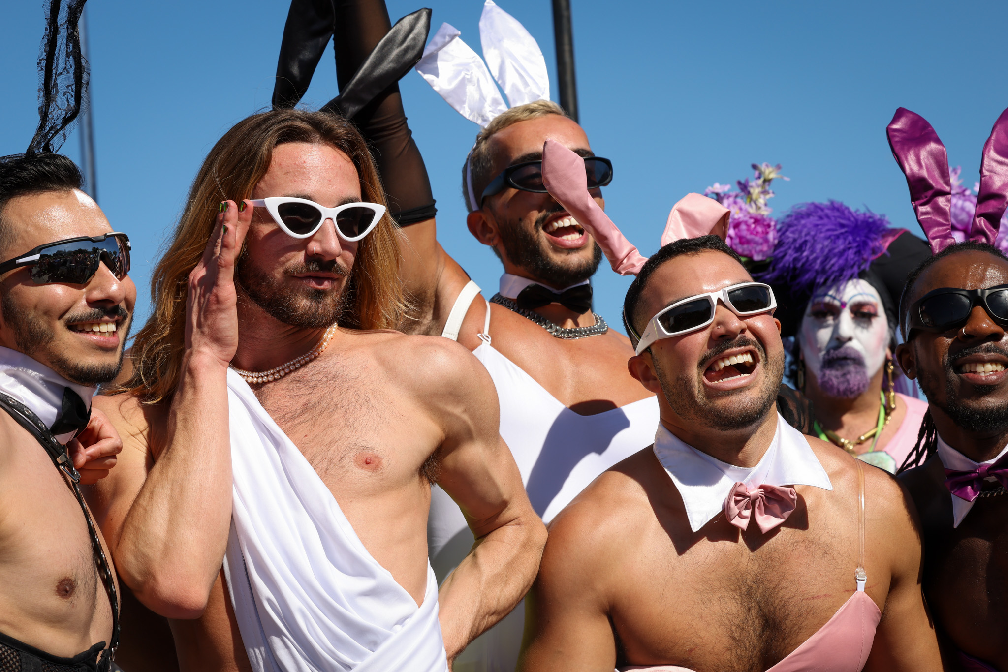 three shirtless smiling men in bunny ears, a toga, a prop collar and/or sunglasses pose looking upward outdoors
