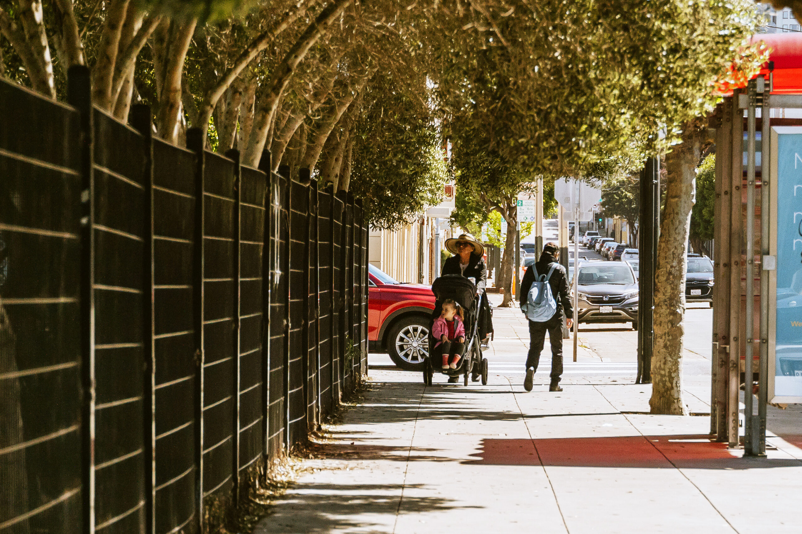 Tree-lined street with a person pushing a stroller and another walking by a black fence.