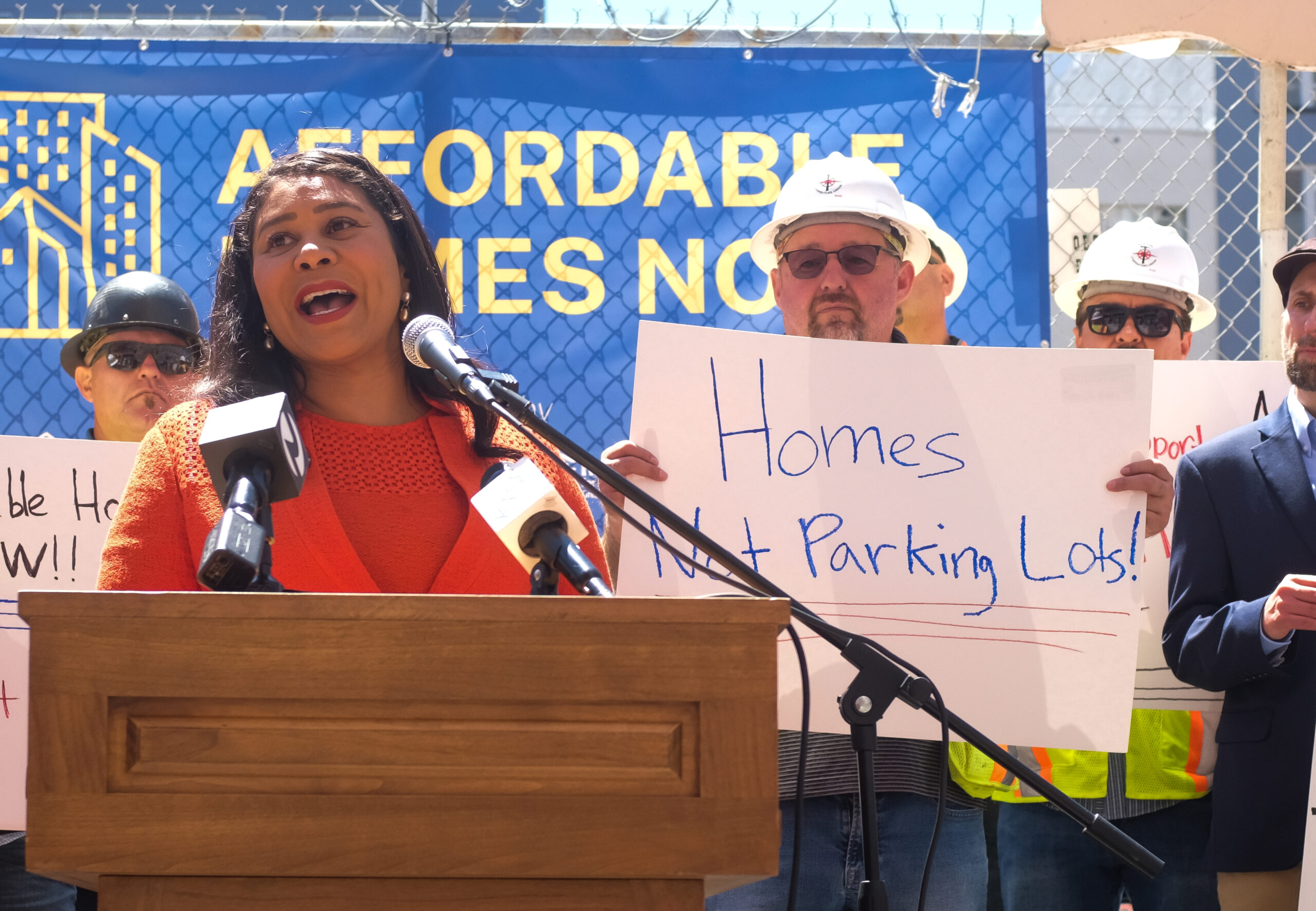 A woman speaks at a podium with microphones, backed by construction workers in helmets holding signs reading "Homes Not Parking Lots." A blue banner reads "Affordable Homes Now."