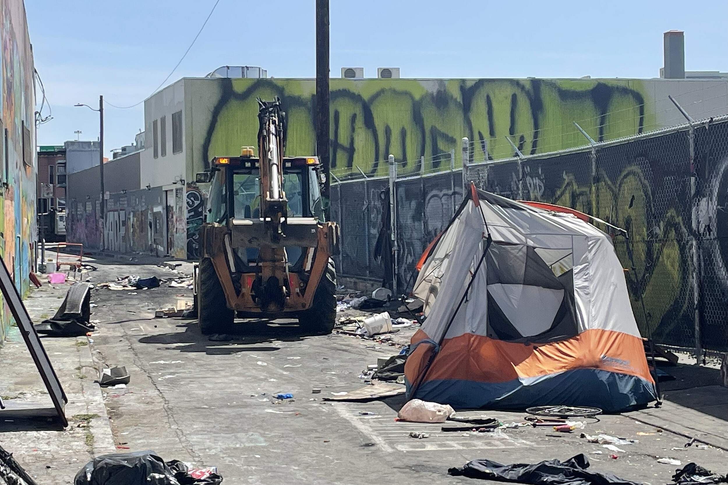 A backhoe near a tent surrounded by litter in a graffiti-covered city alley.
