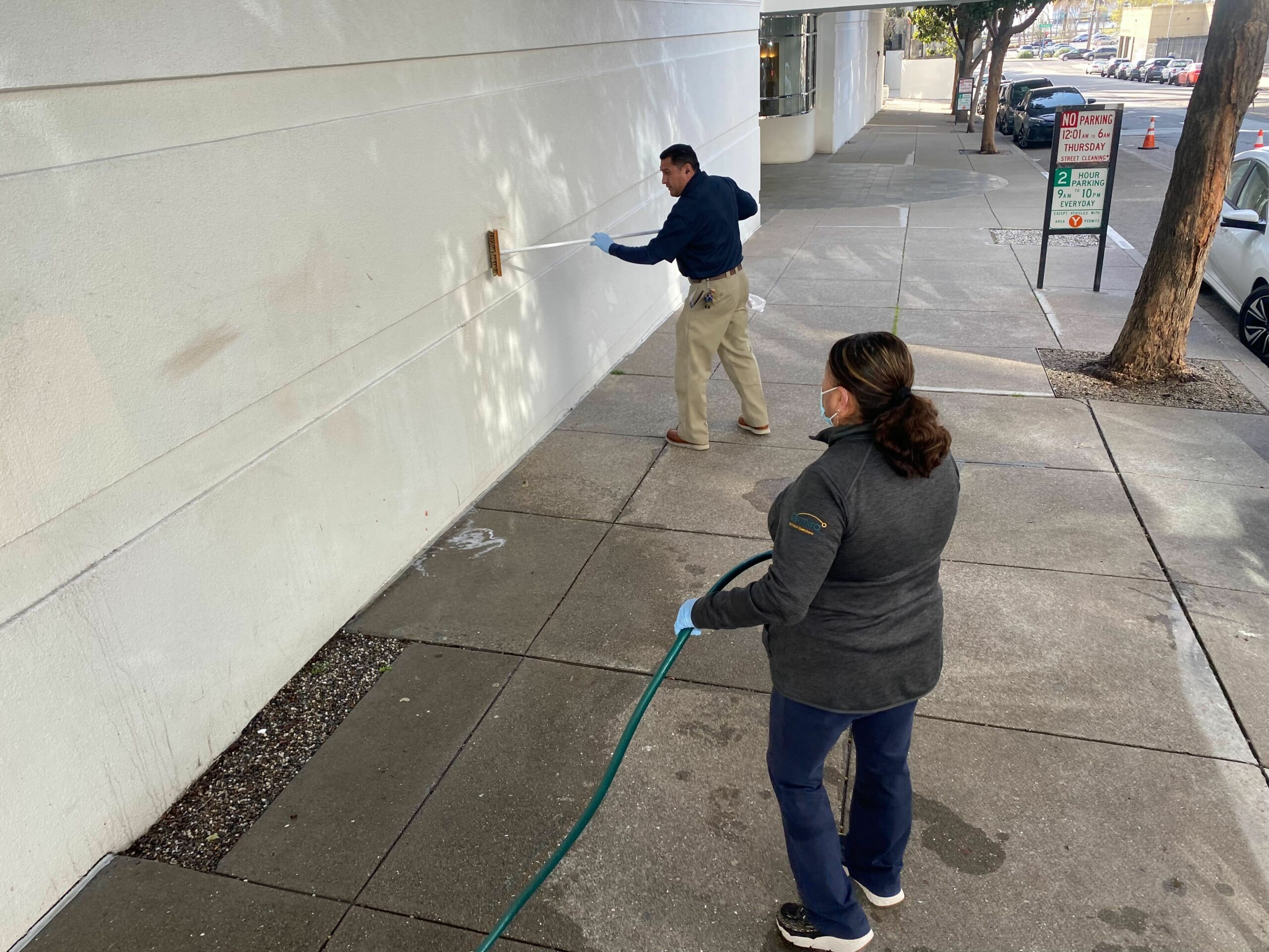 Two people are cleaning a graffiti-covered wall. One person is scrubbing the wall with a brush, while the other holds a hose, wearing gloves and a mask.