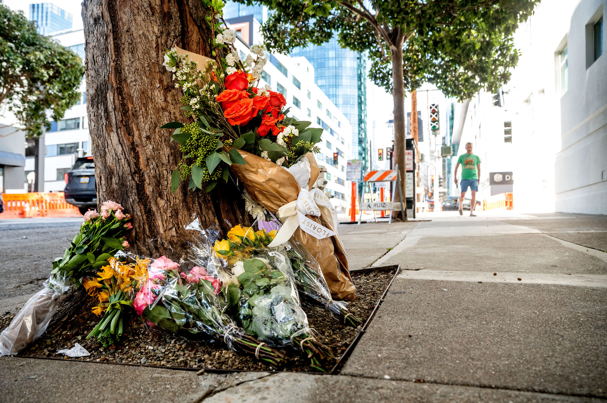 Bouquets of flowers are placed against a tree on a city sidewalk, likely a memorial, with a person walking in the background.