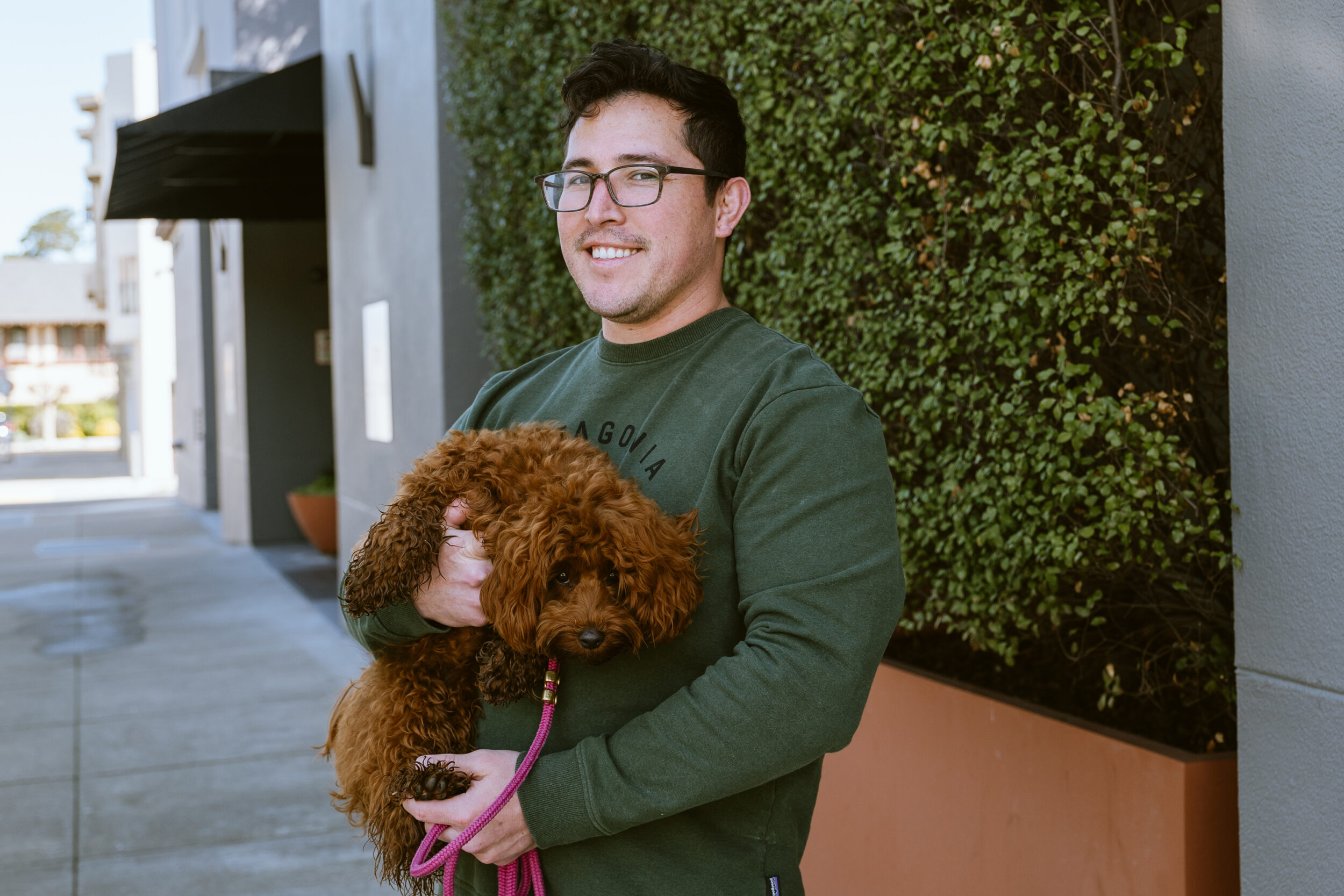 A smiling man in glasses holds a curly-haired dog outdoors near a leafy wall.