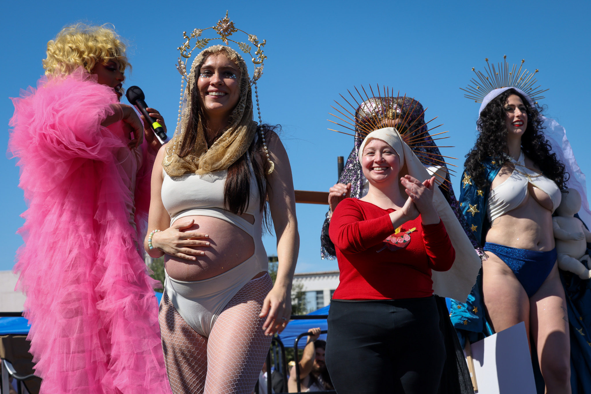 four women or femme-presenting people, one visibly pregnant, stand on a stage outdoors