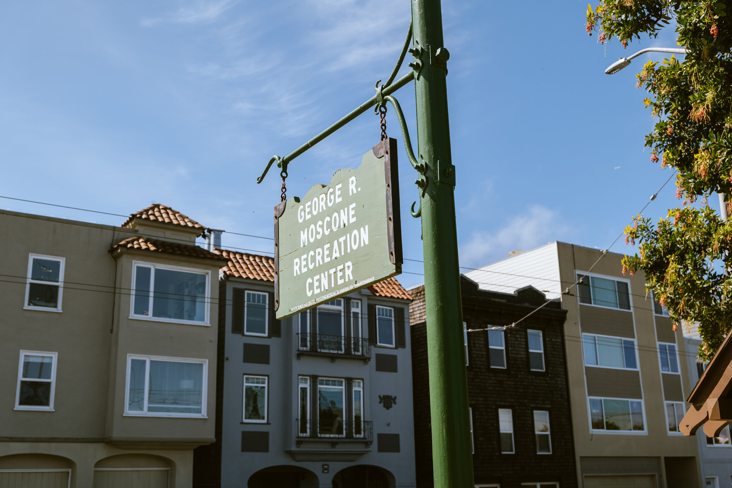 An old, green sign reads &quot;George R. Moscone Recreation Center,&quot; against a backdrop of buildings under a blue sky.