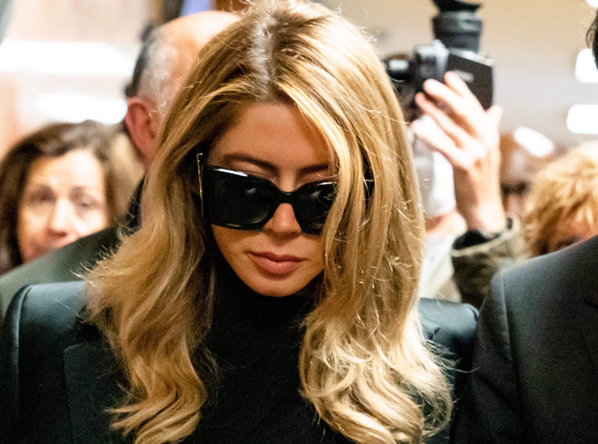 A woman with long blonde hair is wearing large black sunglasses and a black outfit in a crowded setting. People and cameras surround her.