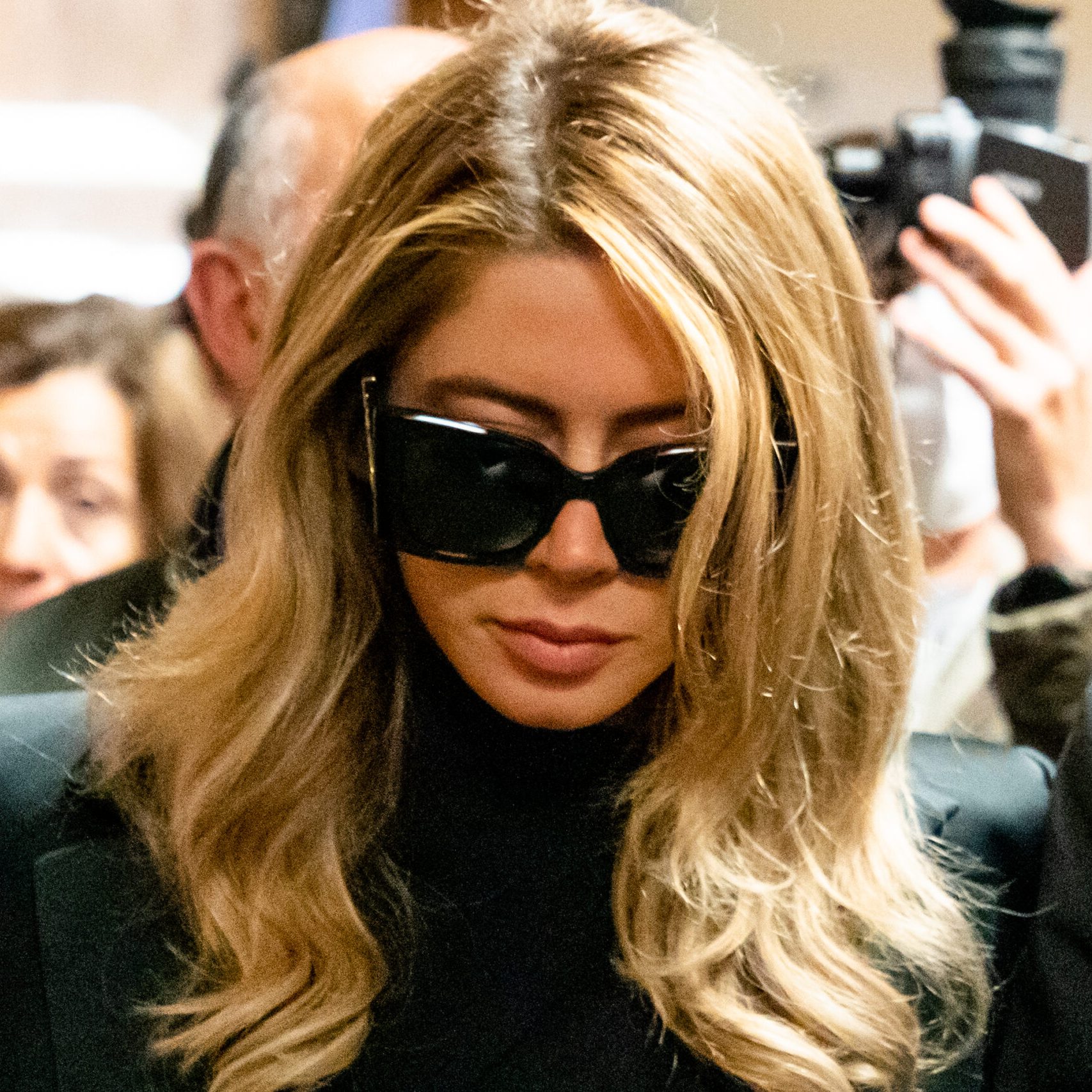 A woman with long blonde hair is wearing large black sunglasses and a black outfit in a crowded setting. People and cameras surround her.