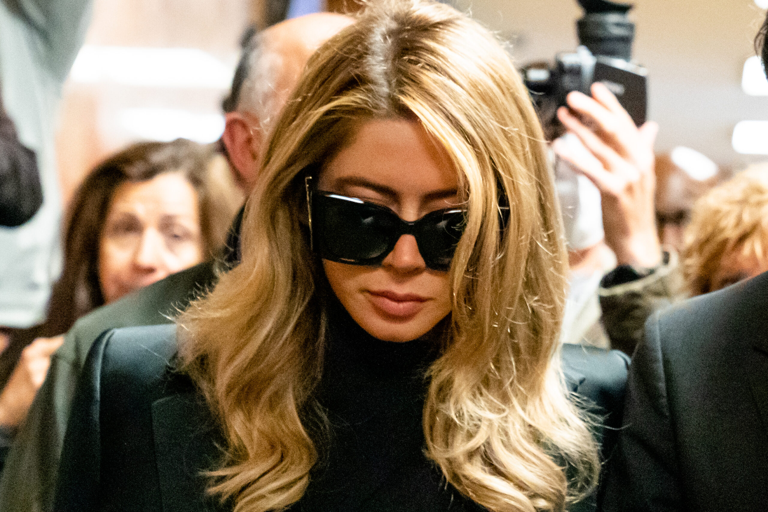 A woman with long blonde hair is wearing large black sunglasses and a black outfit in a crowded setting. People and cameras surround her.