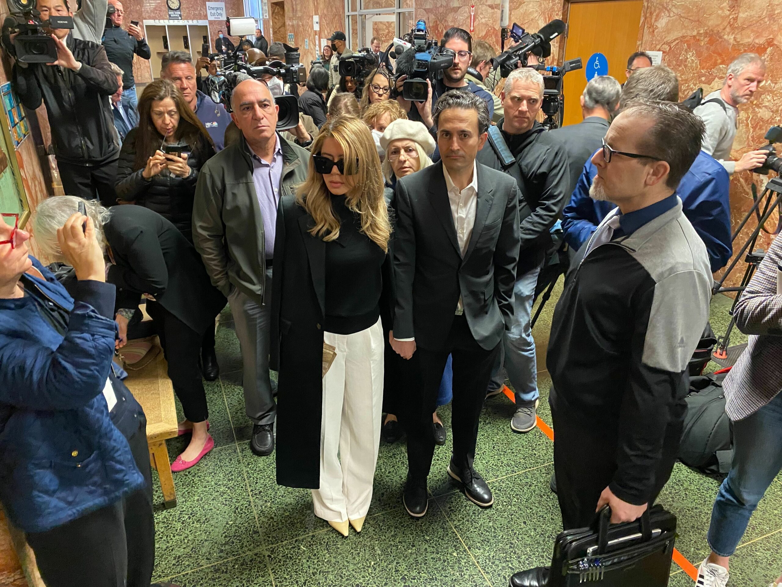 In a crowded hallway, a woman in sunglasses and white pants stands with two men in suits. Cameras and people surround them, capturing the busy scene.