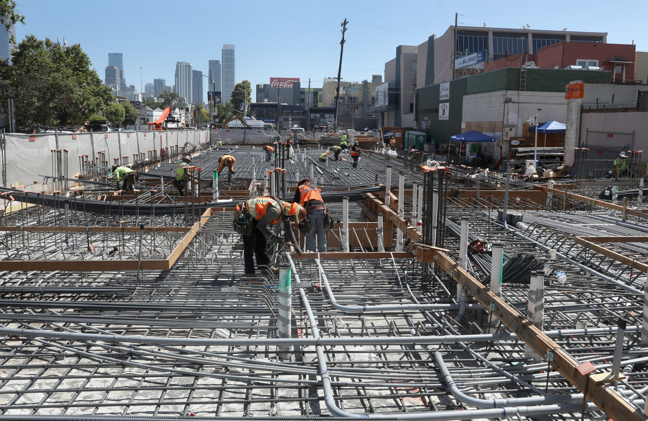 Workers in orange vests are constructing a building foundation with rebar and concrete in a city, with skyscrapers visible in the background.