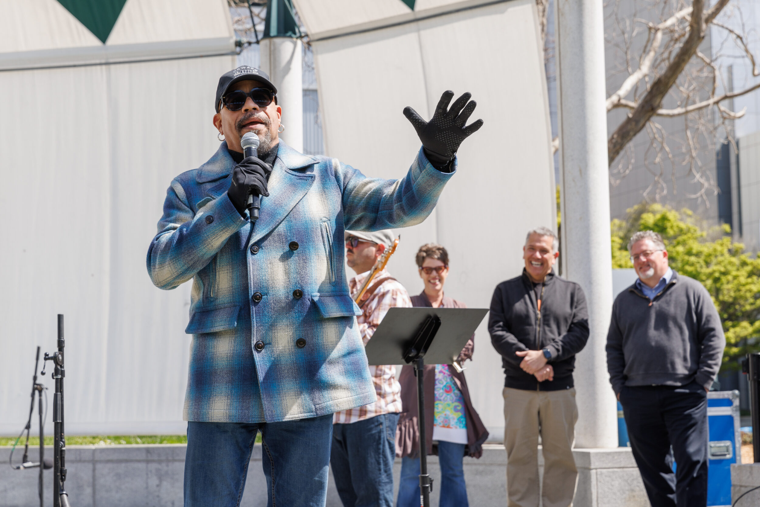 A man in a plaid coat speaks into a mic at an event, raising one hand, with four observers in the background.
