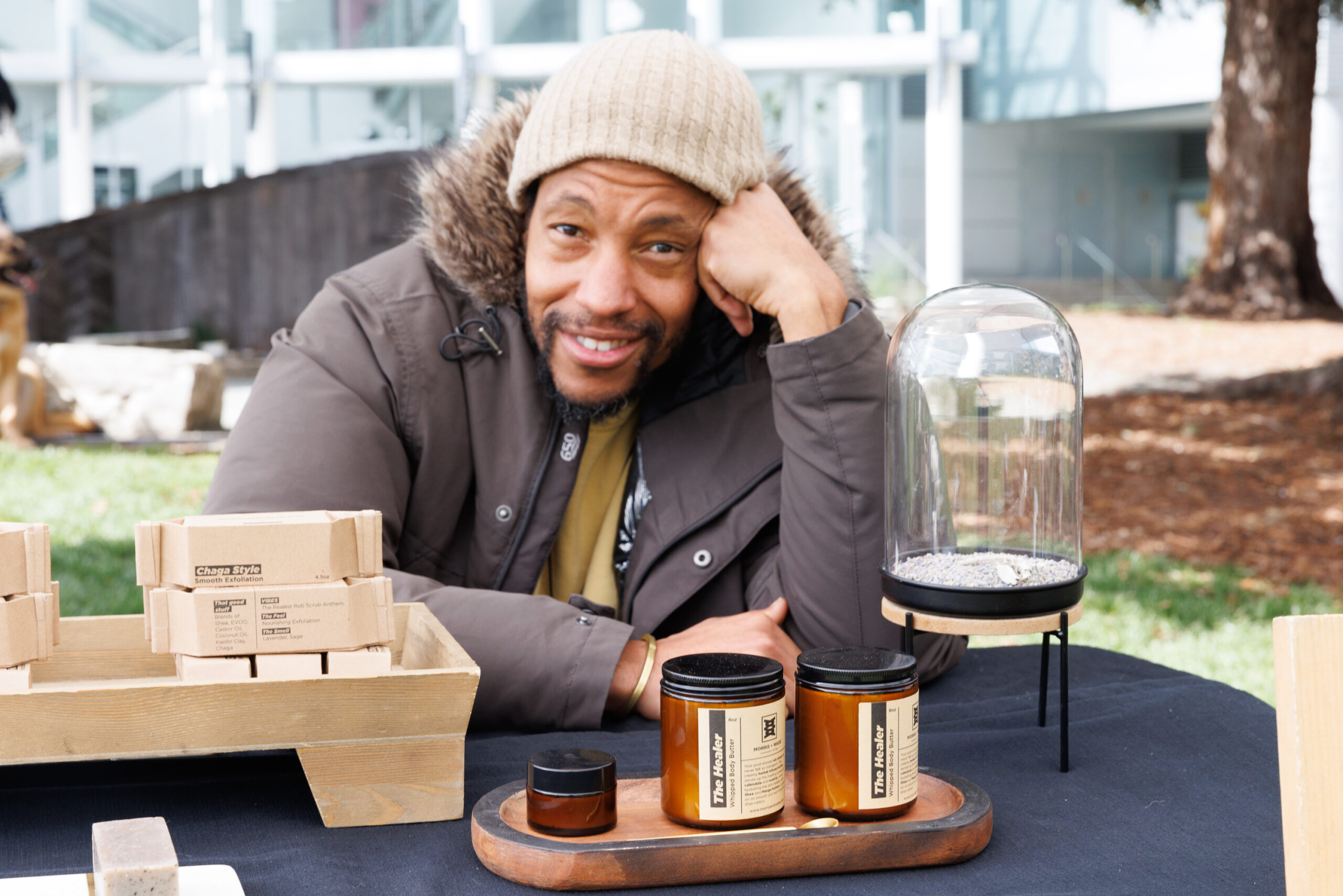 A man smiles at a market stall with candle jars and boxes on display, leaning on his arm, wearing a beanie and jacket.