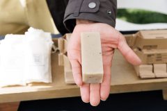 A hand holds a beige bar of soap with packaging materials in the background.