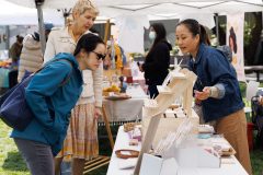 Three women at an outdoor craft market. One vendor explains products to two browsing customers.