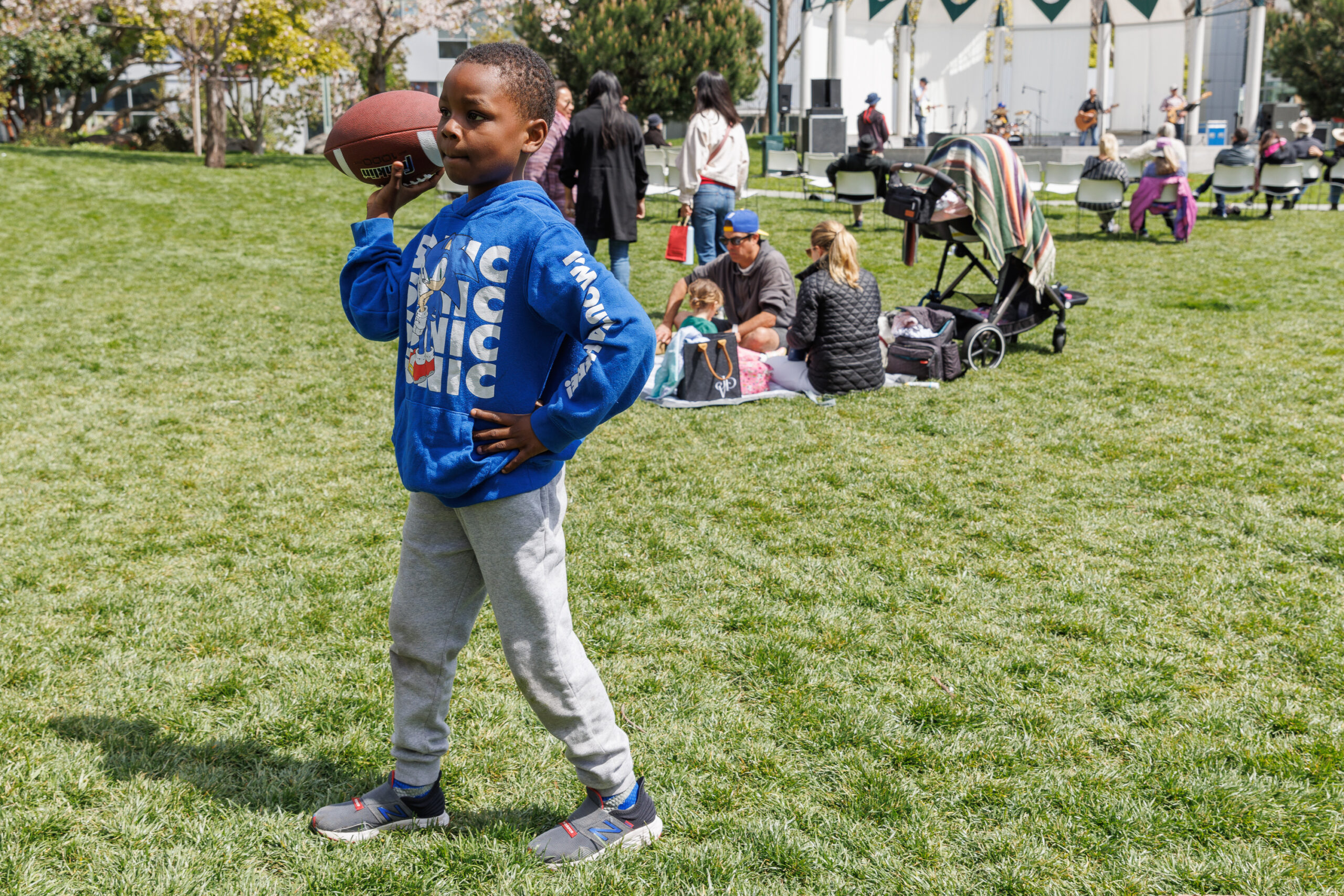 A young African American youth wearing an Sonic the Hedgehog hooded sweatshirt, prepares to throw a football in a large grassy area at Yerba Buena gardens as families enjoy warm weather in the background while a band plays on an elevated stage.