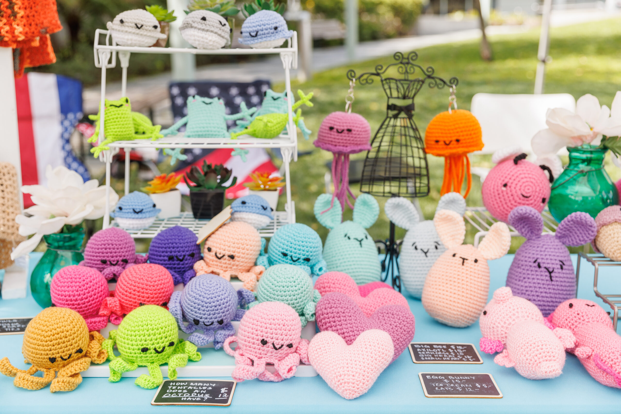 A display of colorful crocheted crafts, including whimsical octopuses, hearts, and bunnies, on a table at an outdoor market.