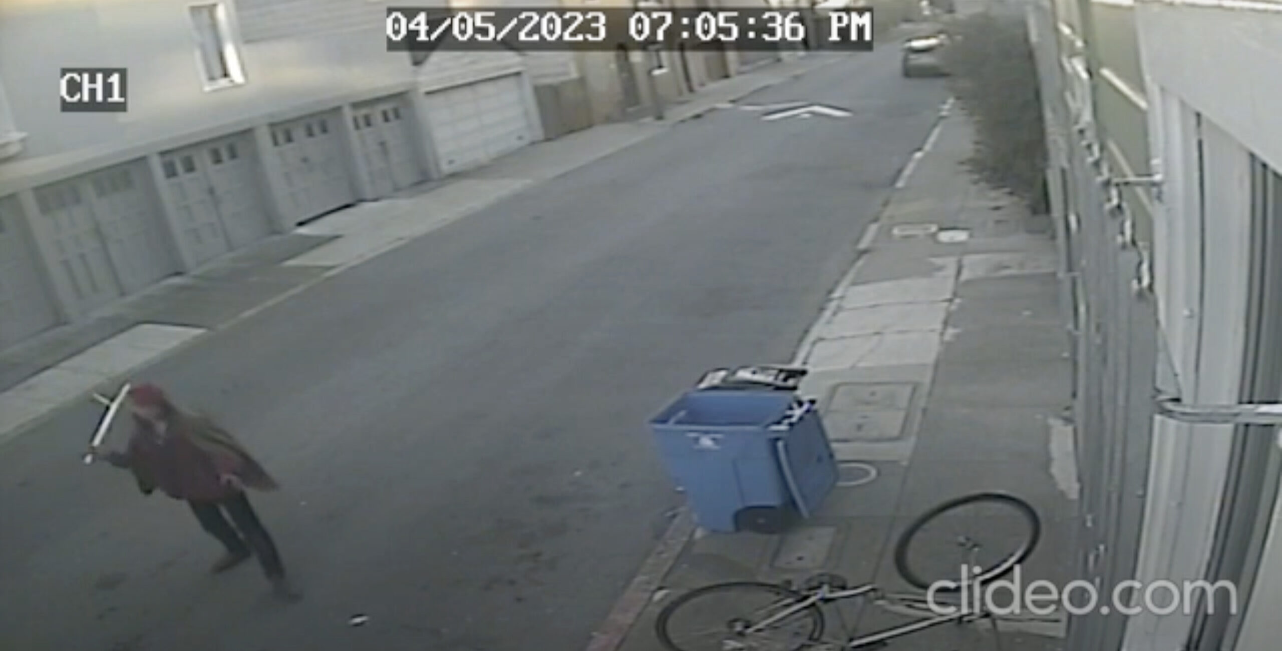 A security camera view showing a person walking past a fallen bicycle and a blue bin on a street.