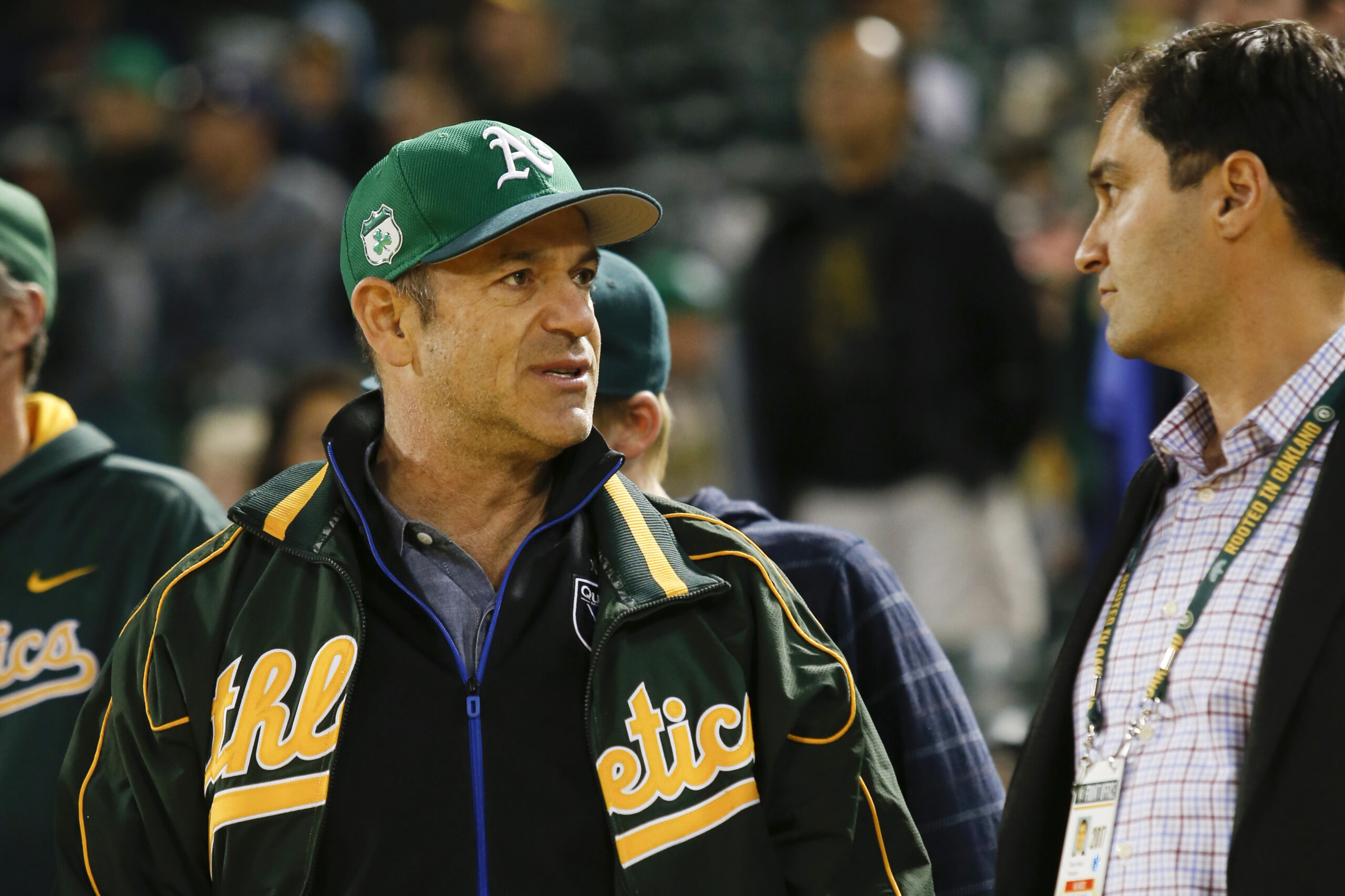 A man in an Oakland Athletics jacket and green Athletics cap stands to the left of a man in a blue shirt.