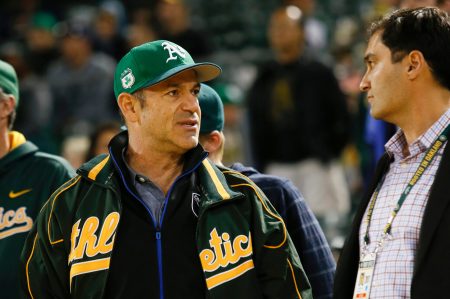 A man in an Oakland Athletics jacket and green Athletics cap stands to the left of a man in a blue shirt.