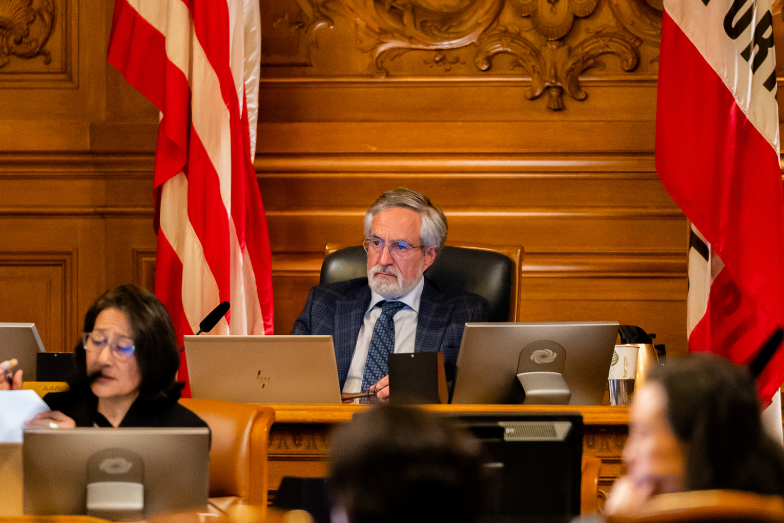 An older man with glasses and a beard sits at a desk, flanked by an American flag and a person working on a laptop.