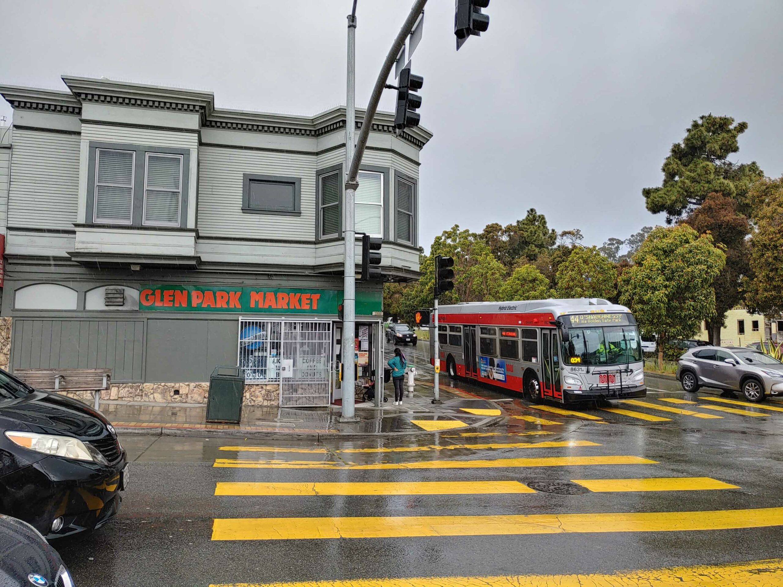 A red bus stops near a crosswalk by the Glen Park Market, on a cloudy and likely rainy day.