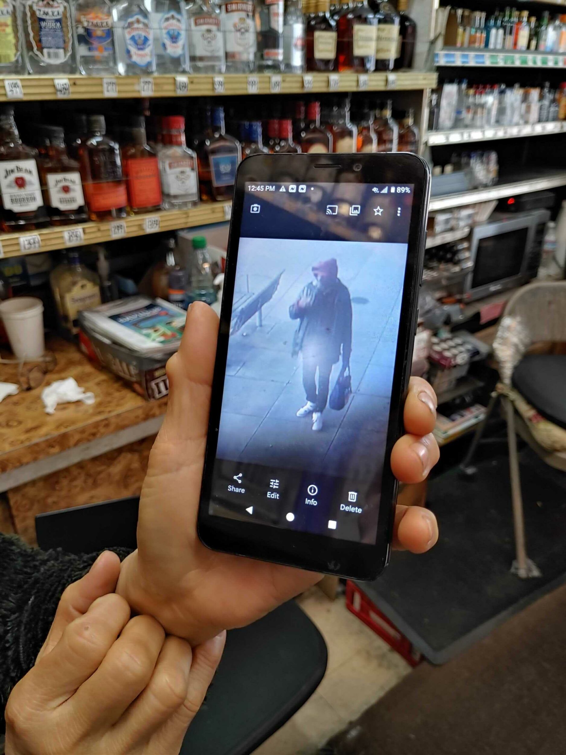 A hand holds a phone displaying a CCTV image of a person, with shelves of bottles in the blurry background.