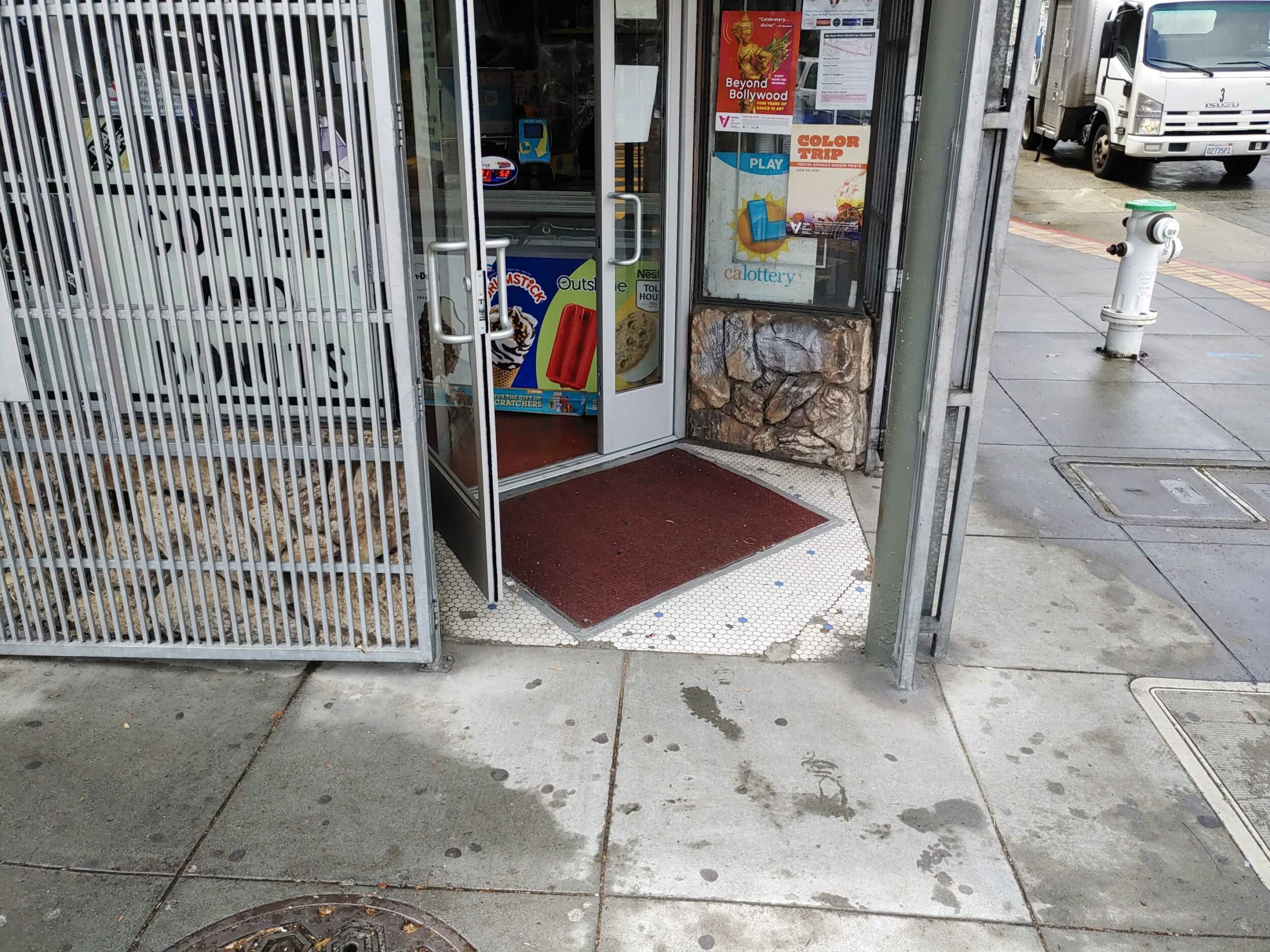 A sidewalk with a storefront; an open security gate reveals ads and a lottery sign on the door.