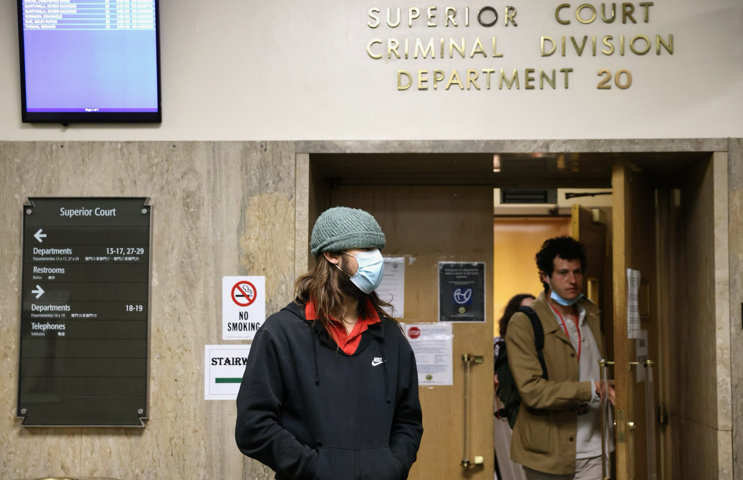 Two people stand near the entrance of a courtroom, one wearing a mask, with signage above and beside them.