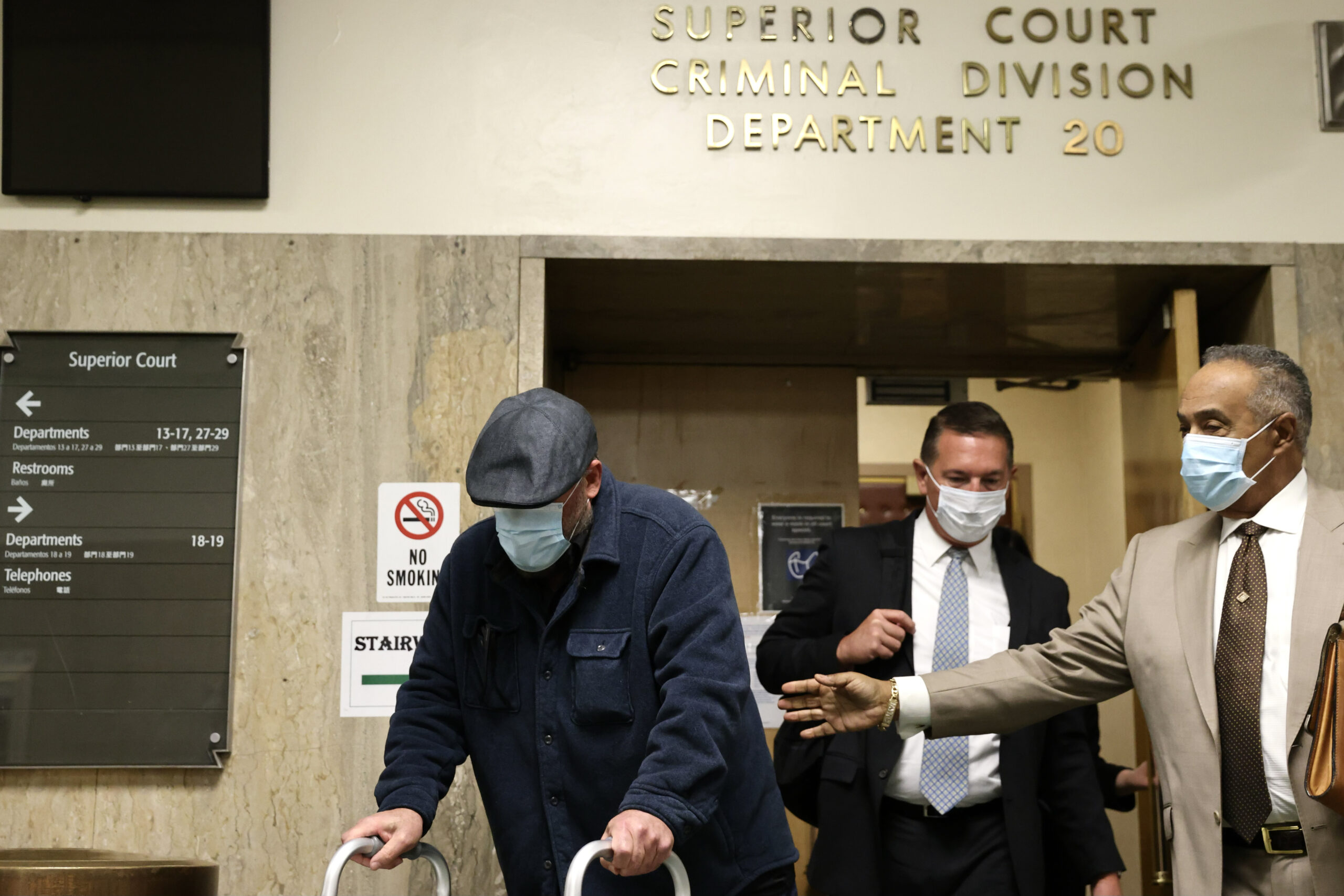 Three men wearing masks walk through a court corridor; one leans on a walker, two appear to assist him.
