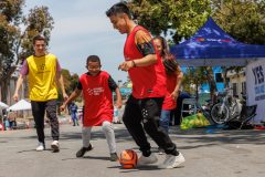 Four people are playing soccer on the street, with a boy in a red shirt dribbling the ball.