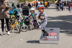 A joyful child in a helmet is playing a bean bag toss game with an iHeartRadio San Francisco board, surrounded by people and bicycles.