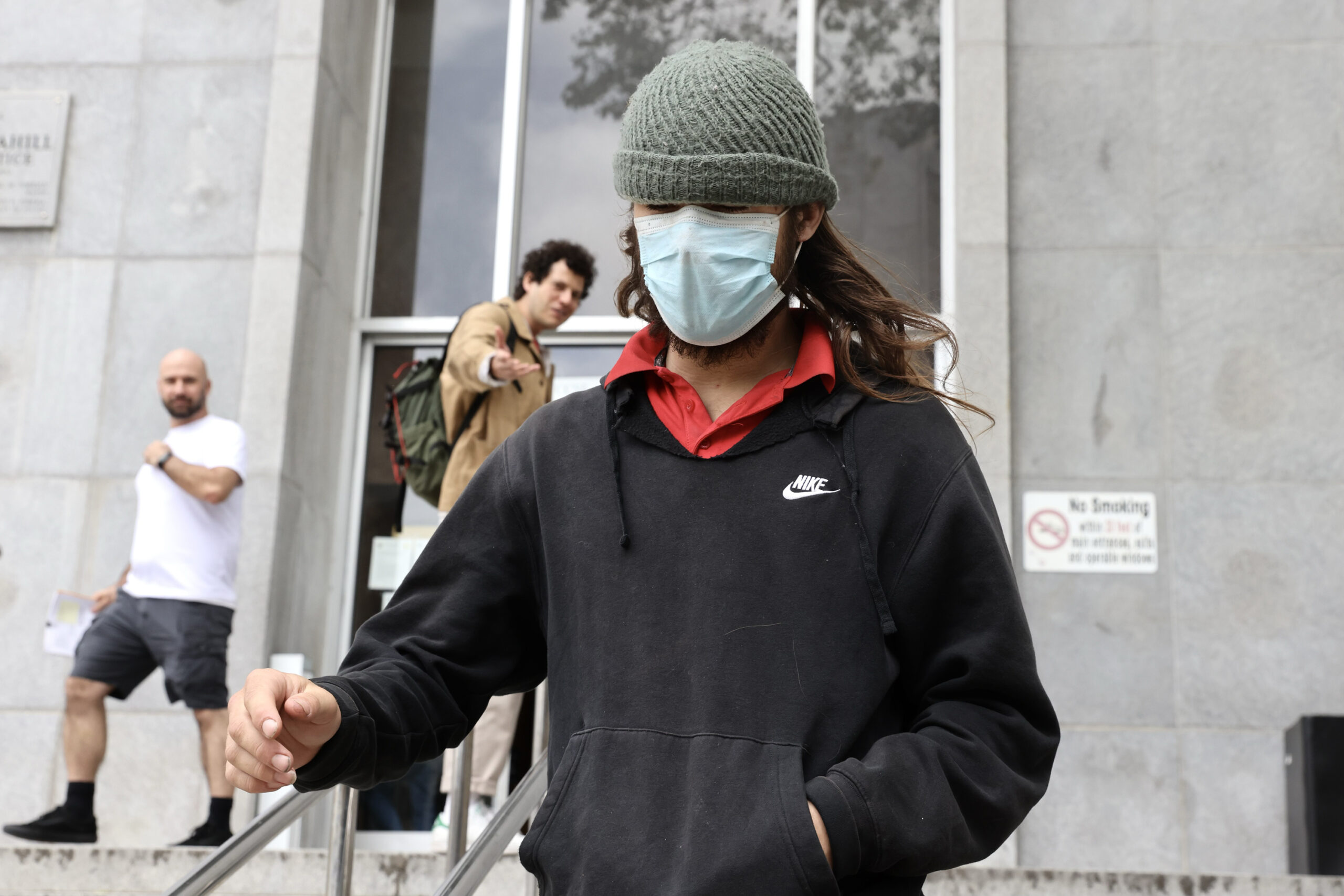 A person in a medical mask, beanie, and Nike hoodie descends steps with two men in the background.