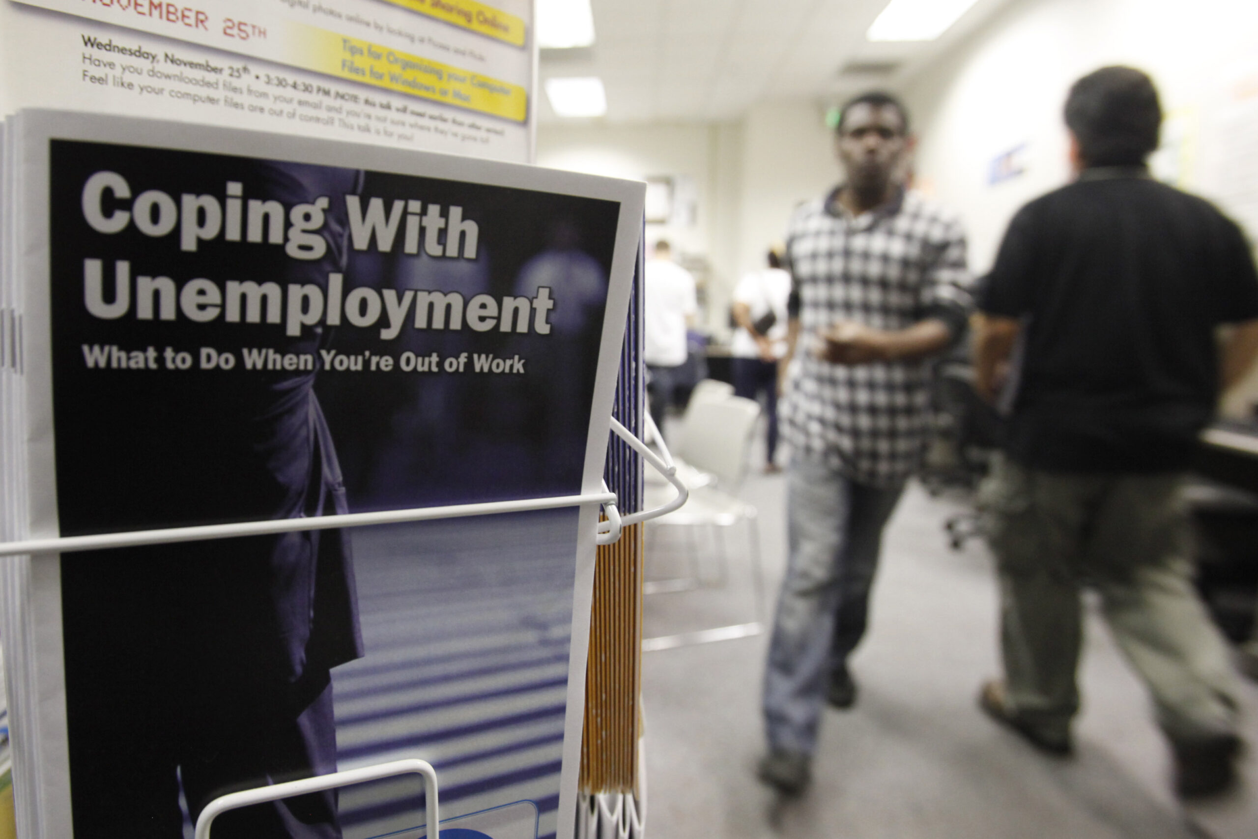 A flyer titled "Coping With Unemployment" in a rack with people blurred in the background.