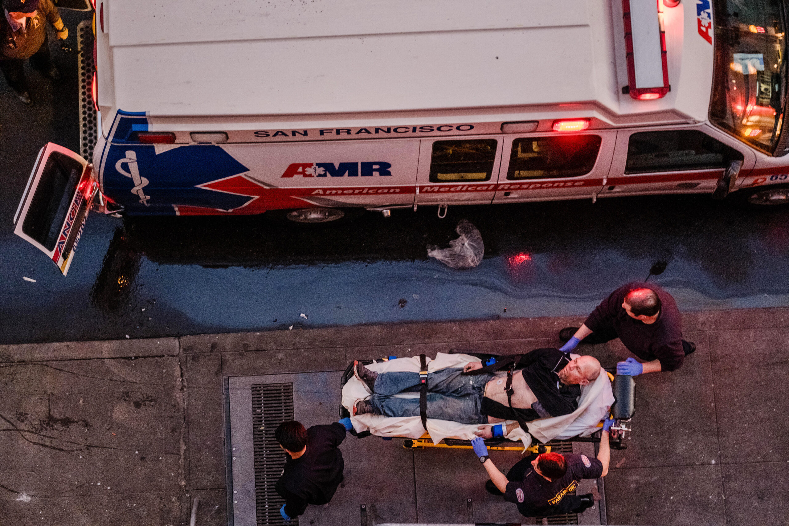 Paramedics attend to a person on a stretcher beside an ambulance on a city street at dusk.