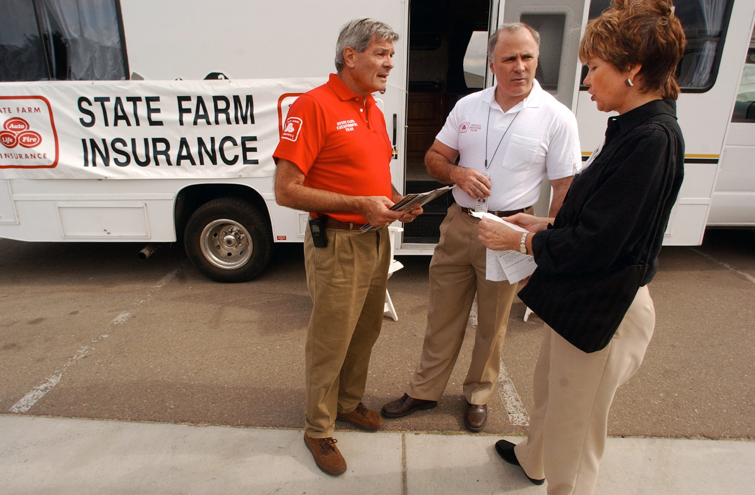 Two men in State Farm insurance shirts speak to woman in front of State Farm vehicle