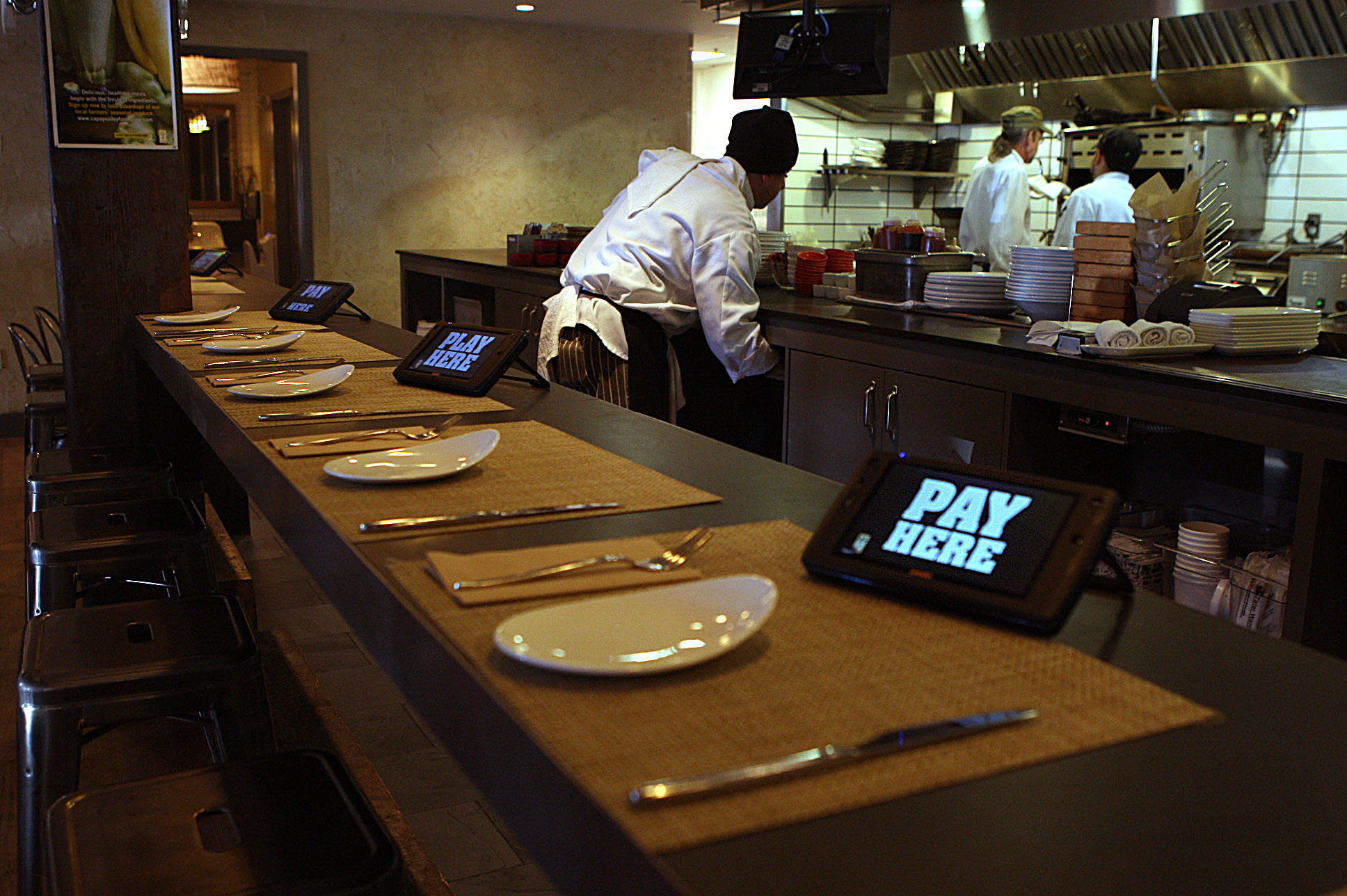 A restaurant counter with place settings of plates and silverware is shown. Tablets displaying "PAY HERE" are placed at intervals. In the background, chefs work in an open kitchen.