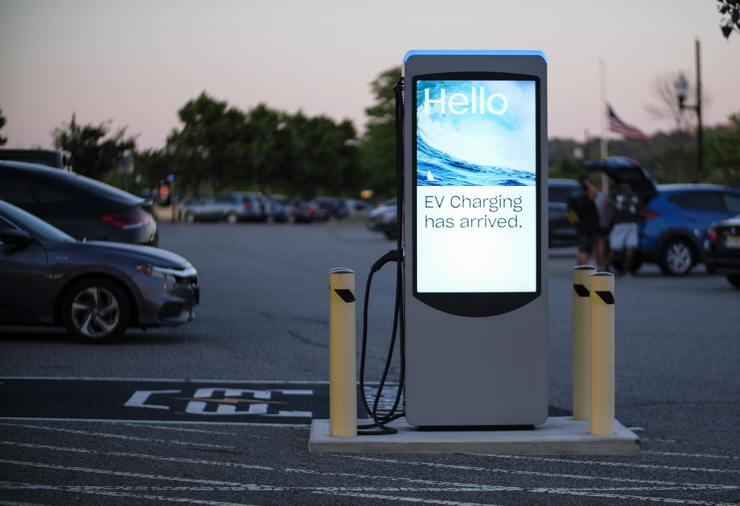 An EV charging station in a parking lot displays &quot;Hello, EV Charging has arrived.&quot; Cars and trees are in the background.